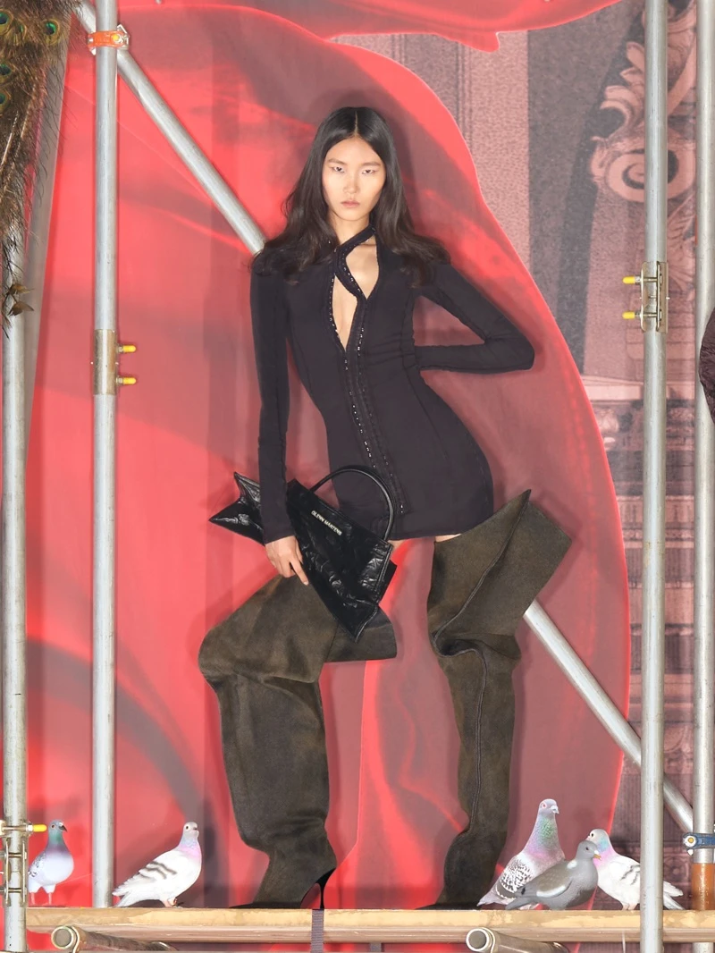 Model in black dress and oversized boots holding handbag on red backdrop