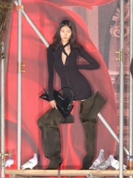 Model in black dress and oversized boots holding handbag on red backdrop