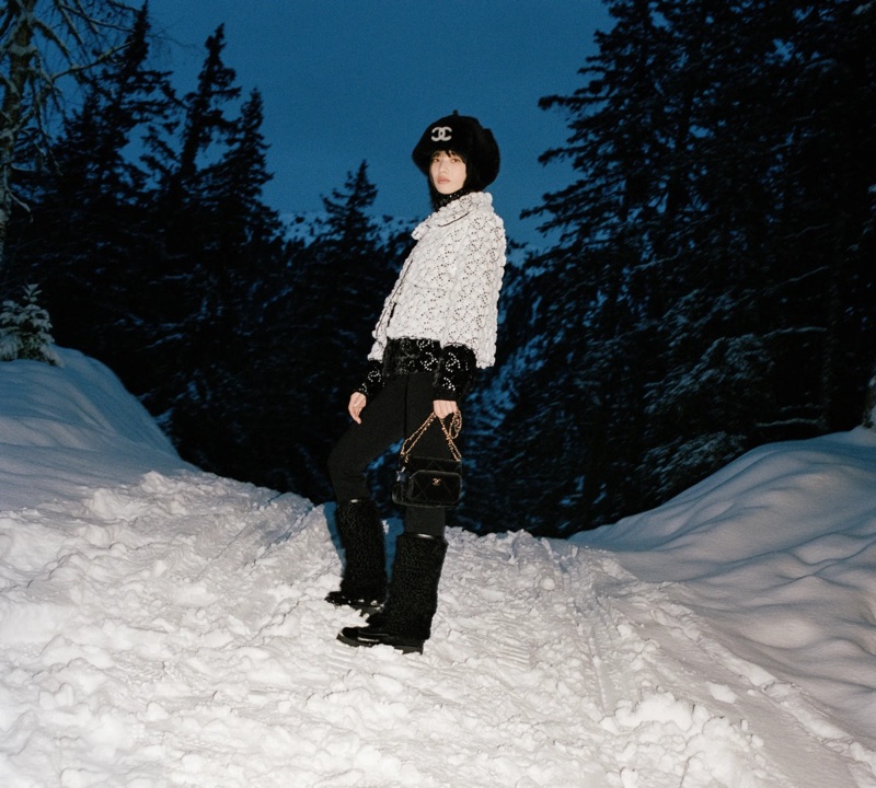 Nana Komatsu in a Chanel textured white jacket, black logo hat, and snow boots standing on a snowy mountain path at twilight.