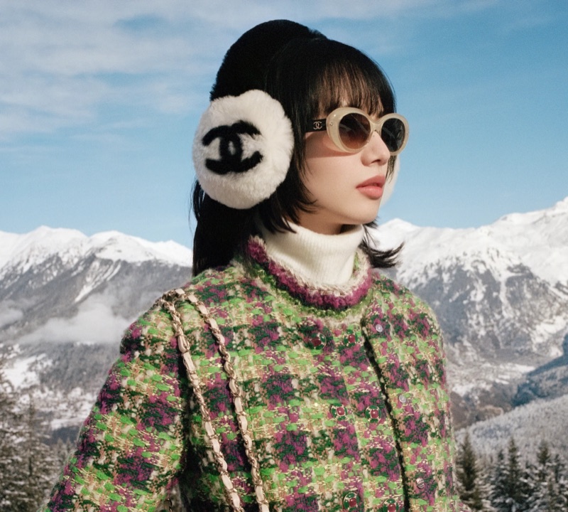 Nana Komatsu wearing Chanel logo earmuffs, sunglasses, and a green tweed jacket against a snowy mountain backdrop.