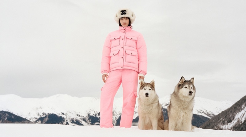 Nana Komatsu wearing a pink Chanel puffer suit and white fur trapper hat standing in snow with two huskies against alpine mountains.