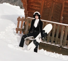 Nana Komatsu in black and white ski apparel holding a snowboard outside a wooden chalet in the snow for Chanel Coco Neige.