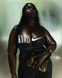 A Black woman with wet hair wearing a dark fitted athletic top holding a small olive green bag against a mottled metallic background.