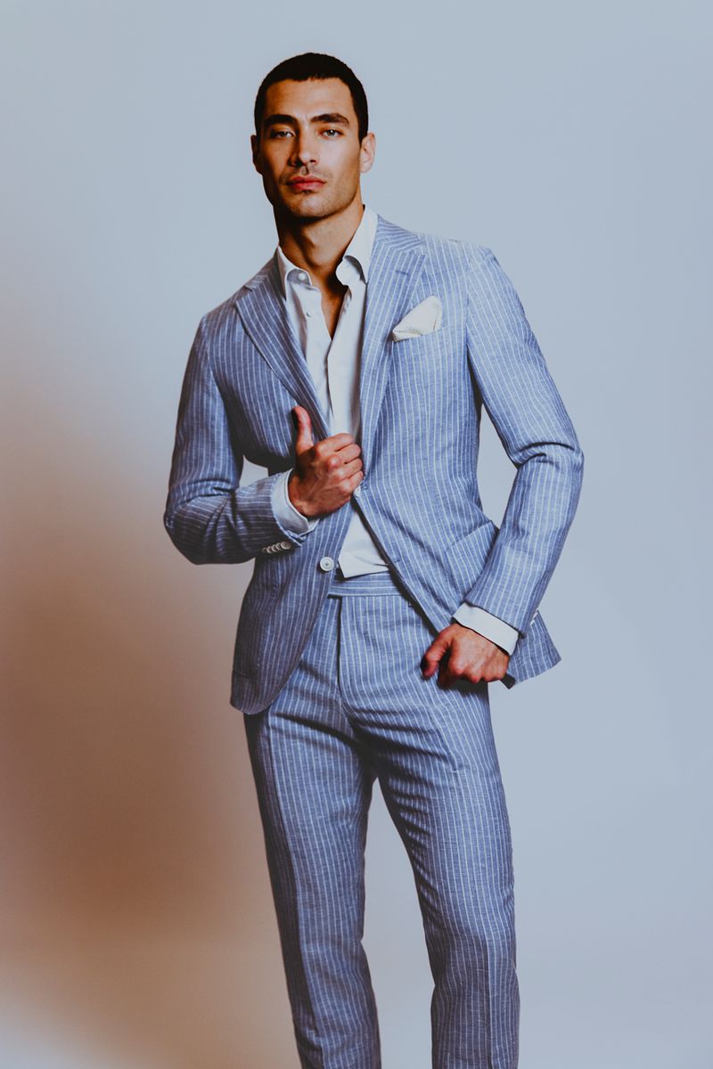 An adult male model poses in a light blue striped suit and white shirt, standing indoors against a light gray background, fashion concept.