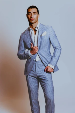 A man posing in a light blue pinstripe tailored suit and unbuttoned white collared shirt against a neutral studio backdrop.