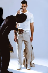 Behind-the-scenes shot of a male model in a white polo shirt and beige trousers being adjusted by crew in a studio.