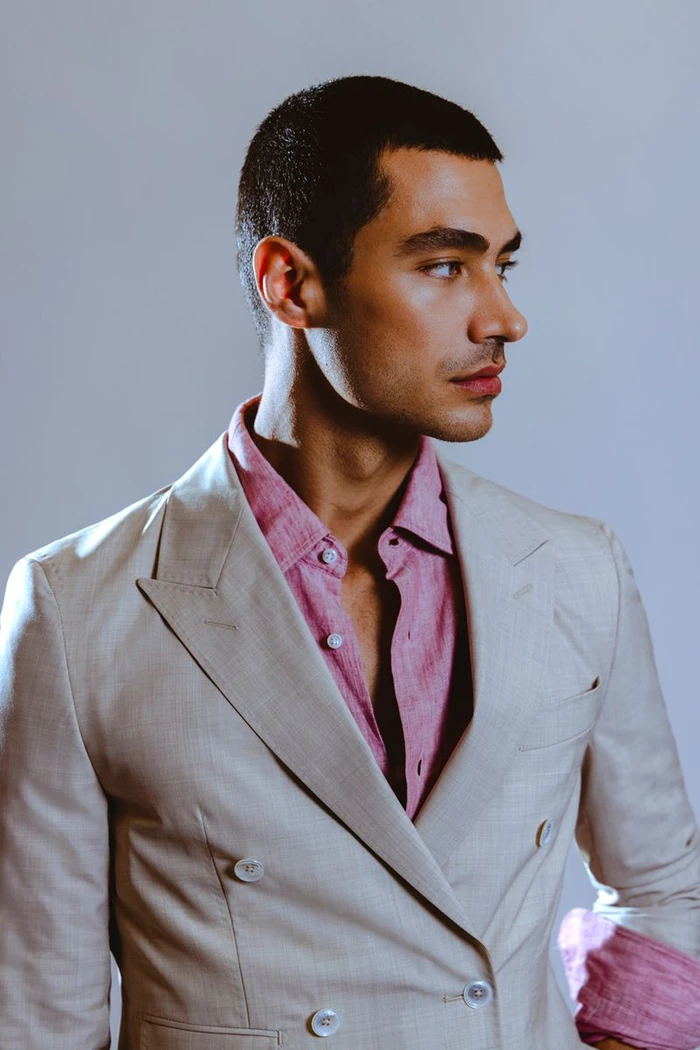 Man with short dark hair in profile wearing a cream double-breasted blazer and pink shirt against a grey seamless background