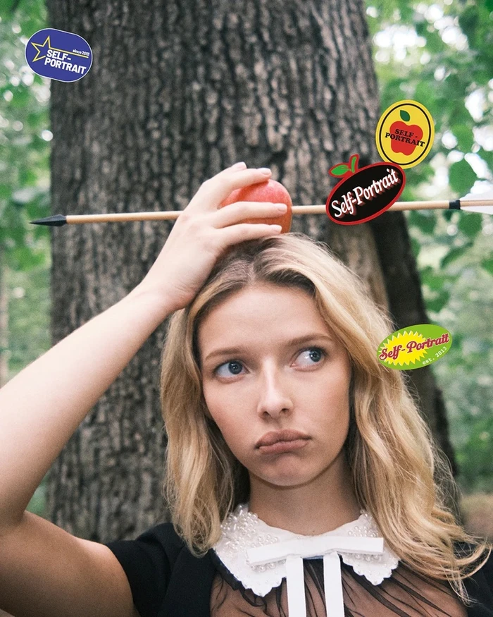 Model Apple Martin balancing an apple pierced by an arrow on her head in a woodland setting for Self-Portrait Fall-Winter 2025 campaign.