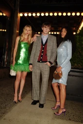 Three models in festive evening wear, including a green feather mini dress, plaid suit, and blue sequin dress, pose on a sidewalk at night.
