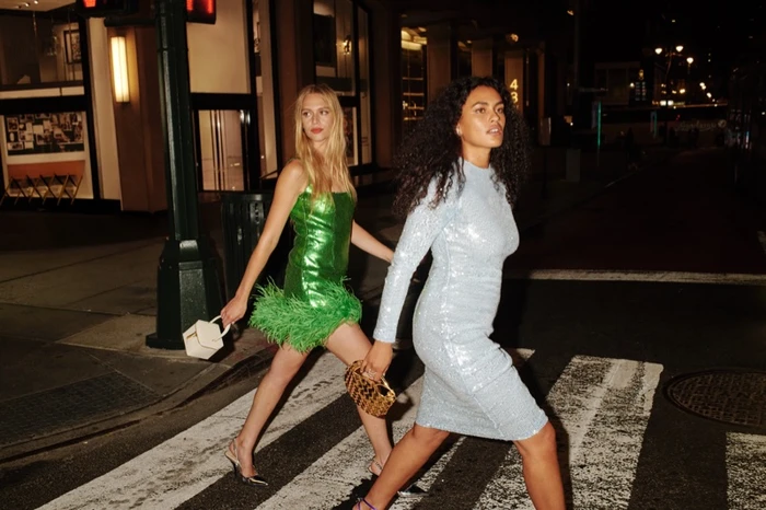 Two women walking across a city street at night, wearing a green metallic minidress and a light blue sequin dress.