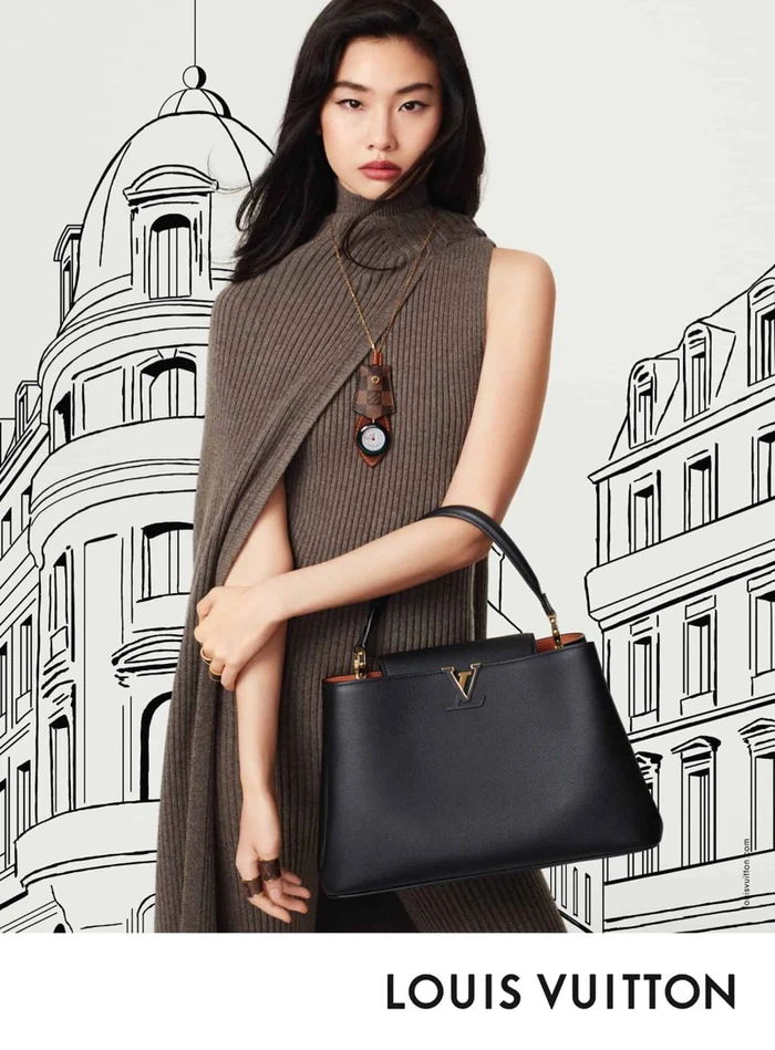 Hoyeon Jung in a brown sleeveless turtleneck holding a black leather handbag against a hand-drawn building backdrop