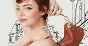 Emma Stone with short auburn hair wearing patterned rings and holding a brown leather handbag against an illustrated backdrop.