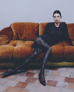 Kendall Jenner lounging on a golden tufted velvet sofa wearing black lace tights and a dark long-sleeve top.