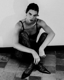 Kendall Jenner in a black bra and patterned lace tights seated on a tiled floor in a monochrome fashion campaign image.