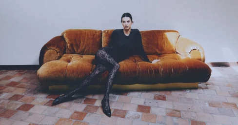 Kendall Jenner lounging on a golden velvet sofa wearing black lace tights and a dark top in an interior setting