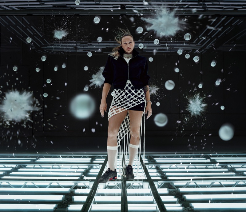 Aryna Sabalenka wearing a black jacket and woven fringe skirt standing on an illuminated light grid floor surrounded by tennis balls.
