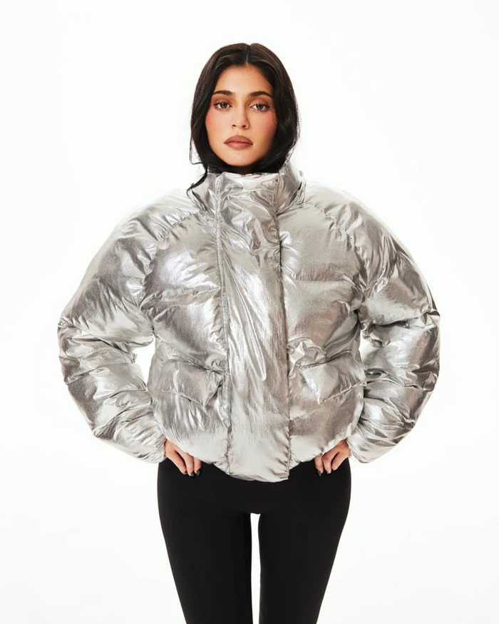 Kylie Jenner wearing a shiny silver puffer jacket and black leggings against a bright white background for Khy.