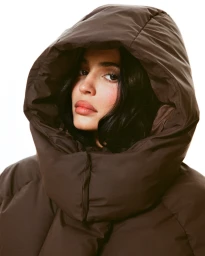 Kylie Jenner wearing a dark brown hooded puffer jacket against a solid white background