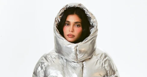 Kylie Jenner wearing a shiny silver metallic puffer jacket with a large hood against a plain white background.