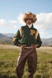 Model in green varsity jacket and fur hat standing in a grassy meadow with distant mountains under a blue sky.