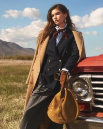 Model leaning against a red truck wearing a camel coat, navy blazer, and holding a suede handbag in a field