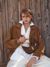 Model in brown fringe suede jacket and white shirt holding a shoulder bag against a rustic wooden wall