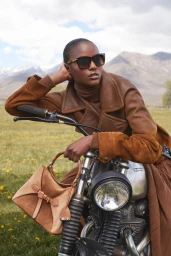 Model wearing brown jacket and sunglasses leaning on a motorcycle with a matching handbag outdoors in a landscape