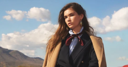 Model wearing a dark blazer, light blue shirt, burgundy neck scarf, and camel coat against a mountain and sky background