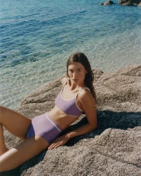 Model Grace Elizabeth wearing a purple bikini on rocky coast by the ocean for Erès
