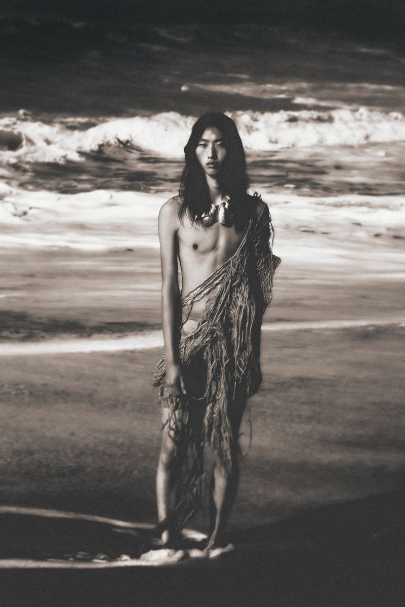 Dramatic black and white portrait of an adult male model with long hair and a shell necklace on a beach with ocean waves.