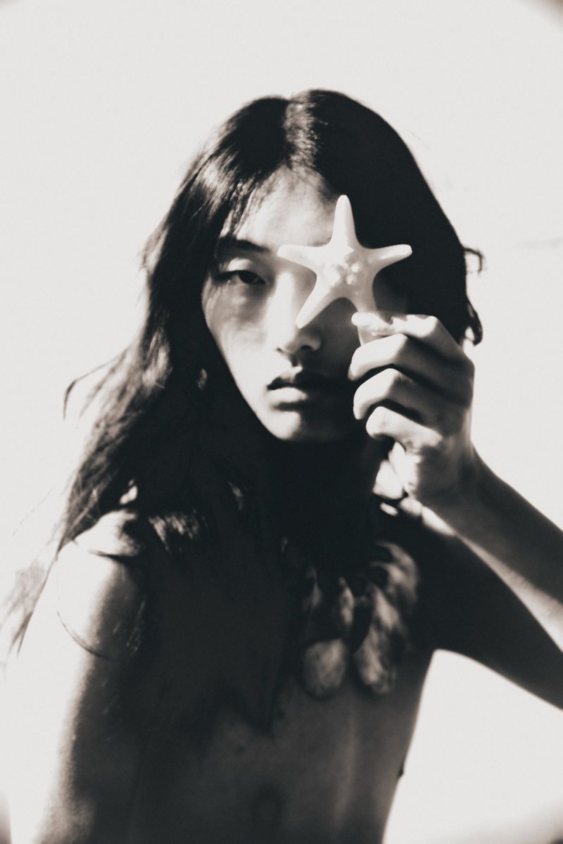 Monochrome portrait of an Asian woman holding a starfish over her eye, with long dark hair and minimal clothing against a white background.