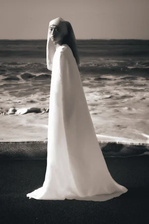 Model wearing a long white hooded cape standing on a dark sand beach with ocean waves breaking in the background.