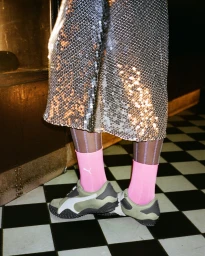 Amelia Gray wearing olive Puma Mostro sneakers with pink socks and a sparkling sequin skirt on a checkered floor.