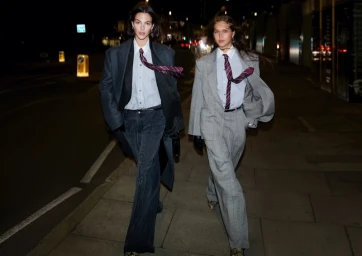 Two models walking down a dark city street at night, wearing oversized grey suits, white button-down shirts, and striped ties.