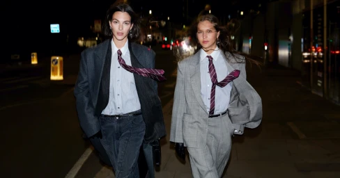 Two female models walking outdoors at night wearing oversized grey suits, white button-down shirts, and striped ties.