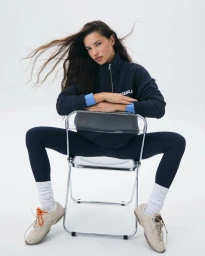 Adriana Lima sitting backward on a transparent chair wearing a navy half-zip sweatshirt and leggings against a white background