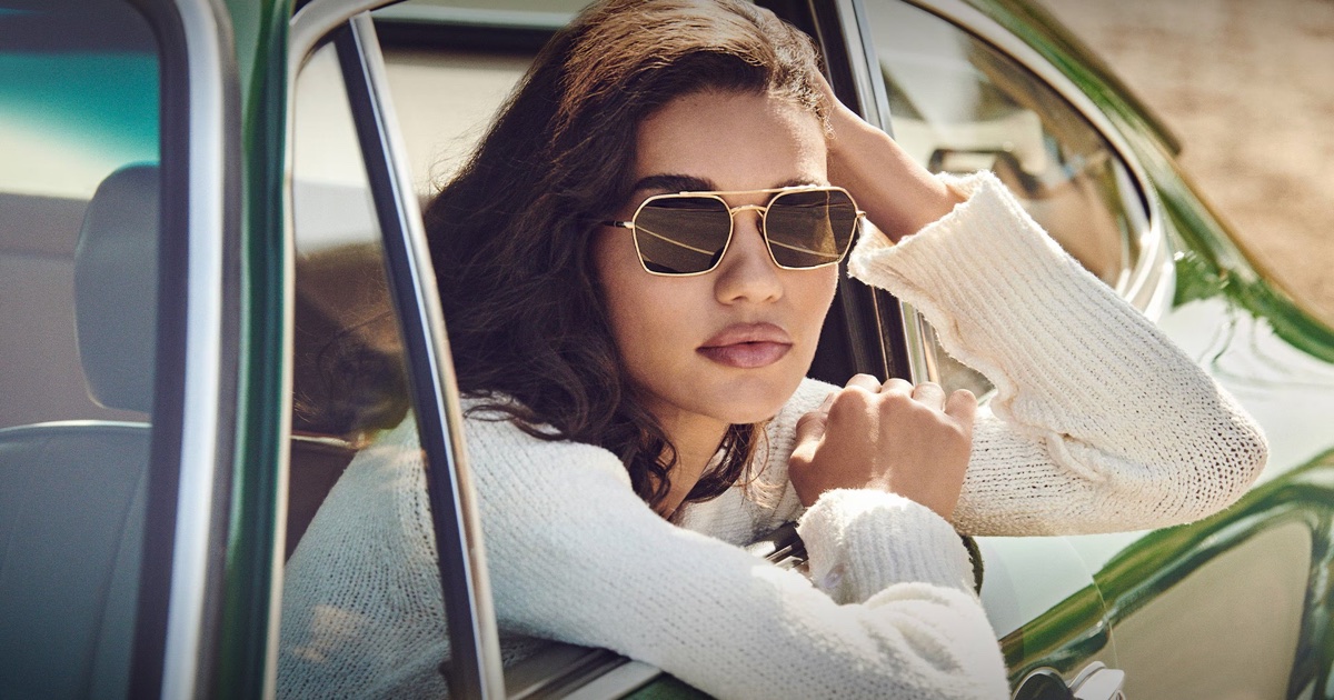 Woman wearing gold hexagonal sunglasses and white knit sweater leaning out the window of a green vintage car.
