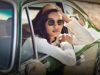 Model wearing gold-framed sunglasses leaning out of the window of a green vintage car for Oliver Peoples Malibu Forever.