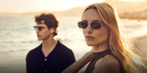 Woman wearing sunglasses in foreground with man standing in background on a sunny beach at sunset