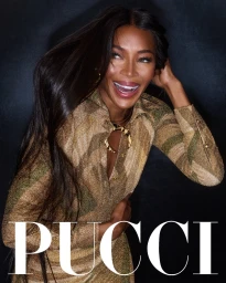 Naomi Campbell smiling in a patterned gold Emilio Pucci shirt against a dark background