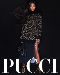 Naomi Campbell posing against a black backdrop wearing a patterned Emilio Pucci turtleneck sweater and holding a black handbag