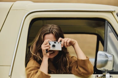 Woman holding a small camera to take a photo while leaning out the window of a pale yellow vintage vehicle.
