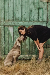 Model Andreea Diaconu leaning down to kiss a Weimaraner dog in front of a weathered green wooden door