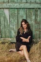 Andreea Diaconu seated outdoors against a weathered green wooden door wearing a dark top and light shoes