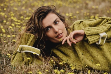 Model Andreea Diaconu lying in a field of yellow wildflowers wearing an olive green turtleneck sweater.