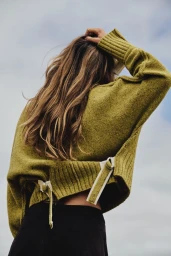 Model from behind wearing an olive green sweater with white strap details against an overcast sky