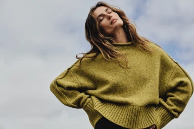 Model with eyes closed wearing an olive green knit sweater against an overcast sky