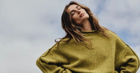 Model wearing an olive green knit sweater posing outdoors against an overcast sky with eyes closed.
