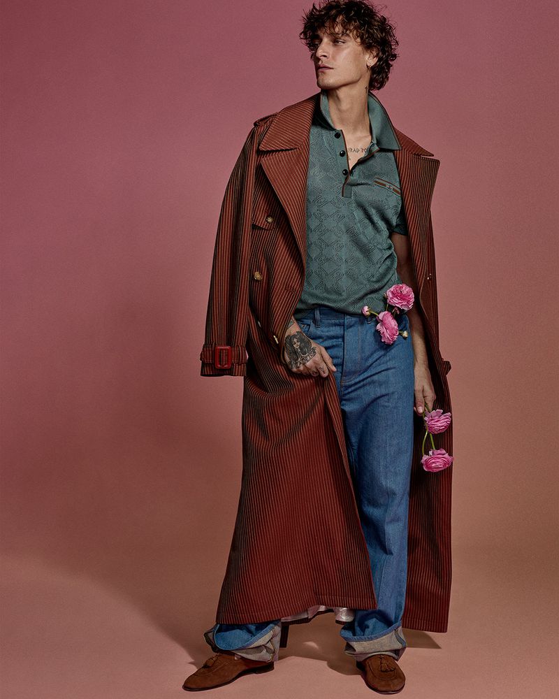 Full-length shot of a young adult male in a brown trench coat, green polo shirt, and blue jeans, holding pink flowers against a pink backdrop.