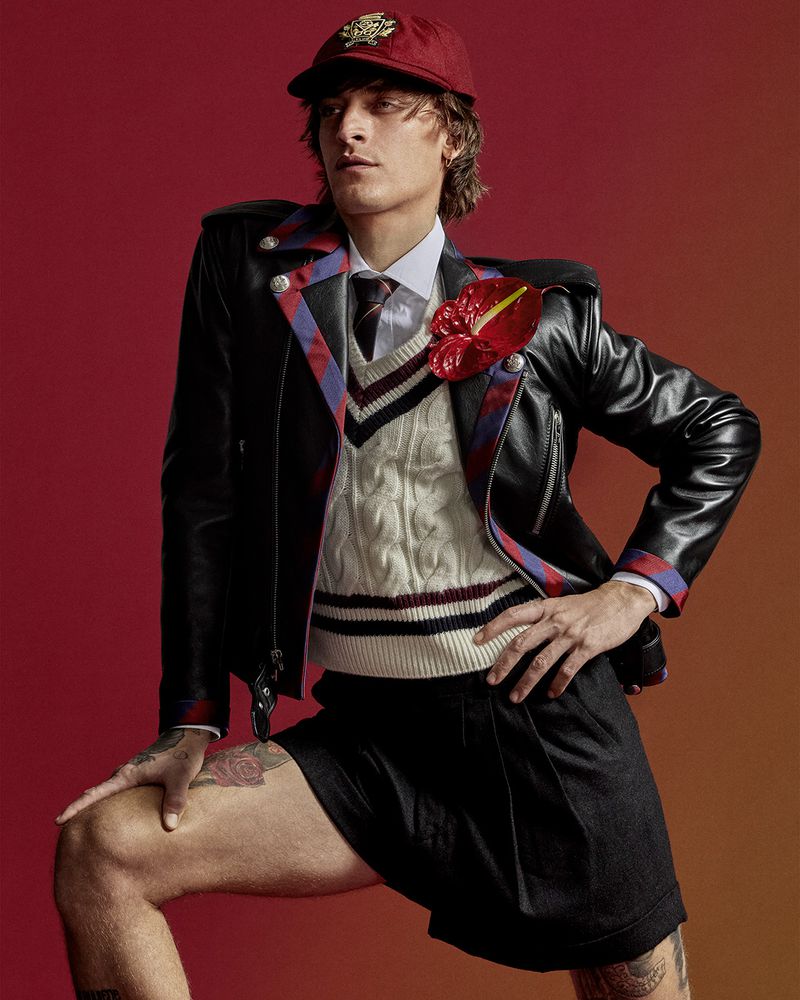 A male fashion model poses with a leather jacket, sweater, cap, and shorts against a studio backdrop with blended red and orange hues.
